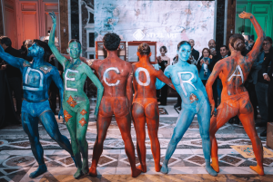 A group of body-painted performers at an artistic event, each displaying a letter on their back to spell "DECORA," symbolizing creativity and transformation in interior design.