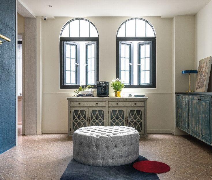 Elegant interior space featuring arched windows, vintage-style cabinetry, a tufted ottoman, and lime-based painted walls.