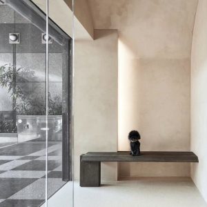 A minimalist interior with lime-based plaster walls, a dark wooden bench, and a small black poodle sitting in a cozy alcove.