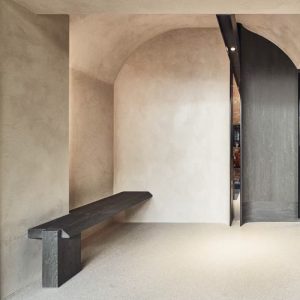 Minimalist interior with arched walls, textured plaster finish, a floating wooden bench, and a curved dark wood door.