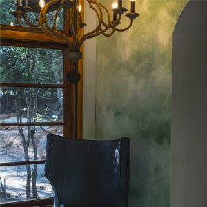 Rustic green lime plaster wall with chandelier and large window overlooking a wooded landscape.