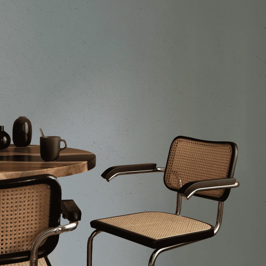 Minimalist table setup with coffee cups, black vases, and cane chairs against a textured blue lime-based painted wall.