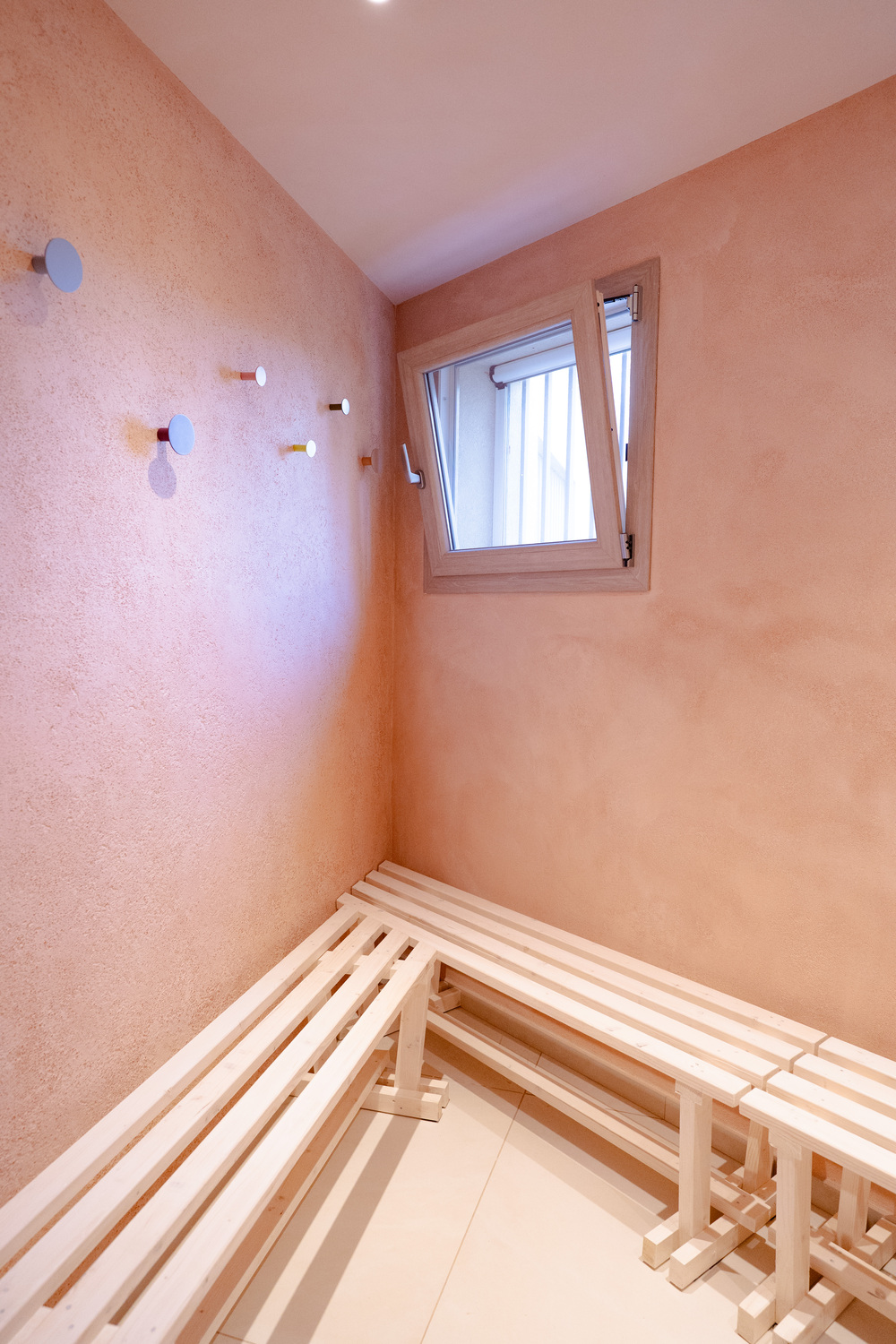 Cocciopesto wall finish in soft peach with modern minimalist wooden benches and colorful wall hooks in a contemporary space.