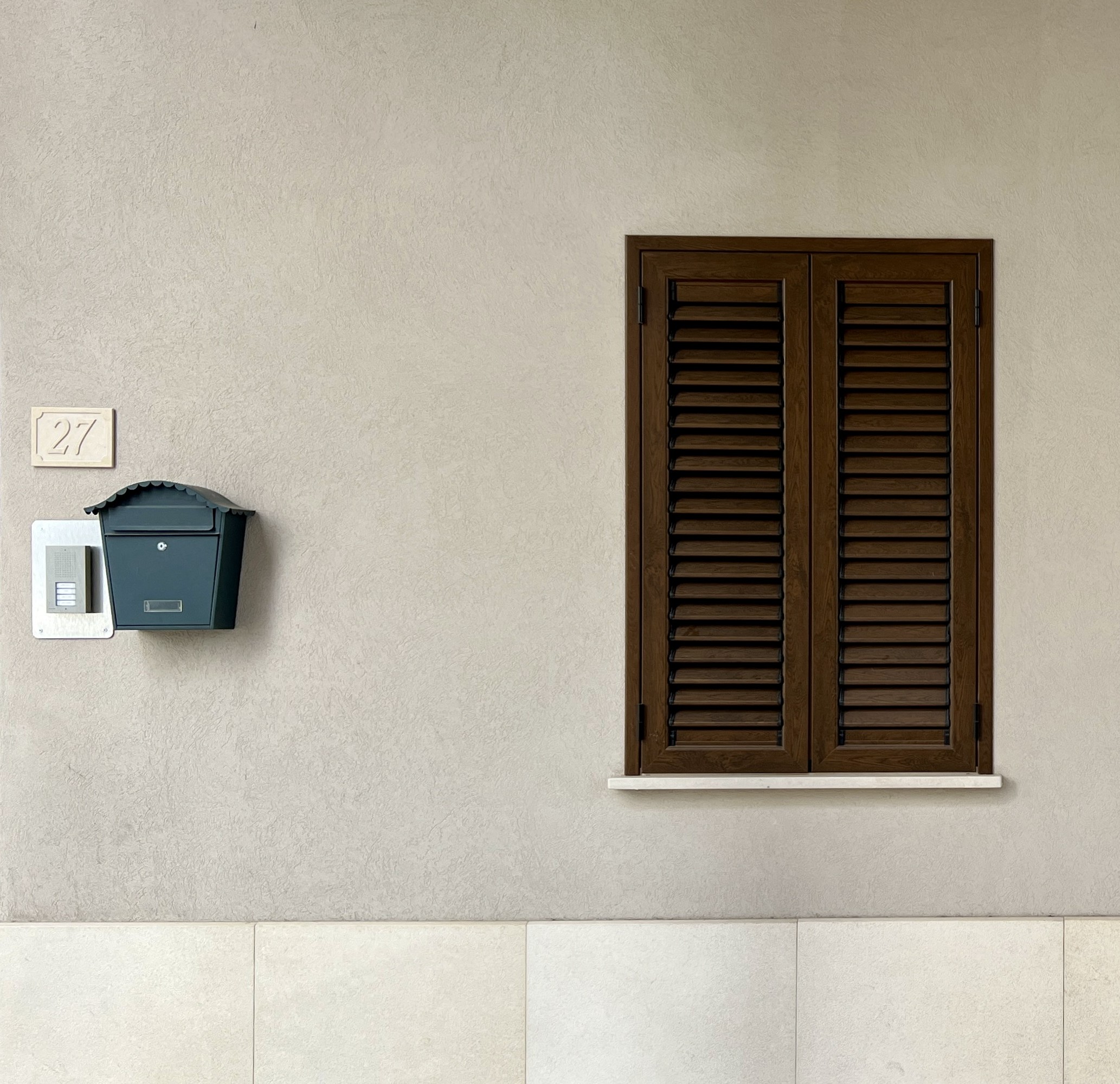 Exterior of a house with a beige wall, brown shuttered window, and a mailbox with the number 27.