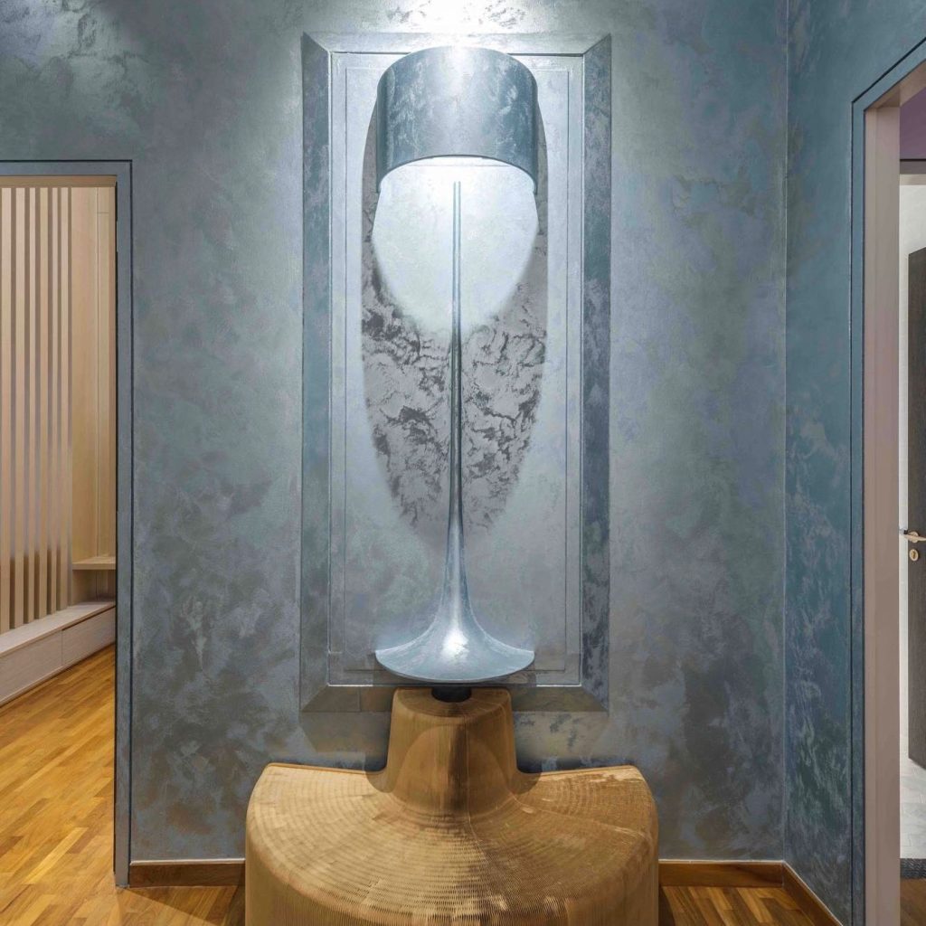 An artistic wall installation featuring luxury metallic wall paint with a 3D textured finish, highlighted by a sculptural lamp design and reflective blue-silver tones.