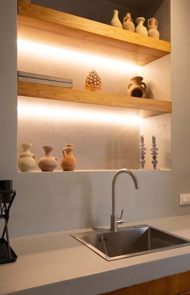 Modern kitchen sink area with matte lime-based plaster walls and wooden open shelving, styled with rustic pottery.