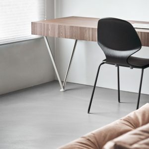 Minimalist home office corner with a wood-grain floating desk, black chair, and light grey microcement floor.