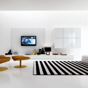 Modern minimalist living room with a white color palette, mustard-yellow lounge chair, black-and-white striped rug, and a built-in bookshelf filled with colorful books.