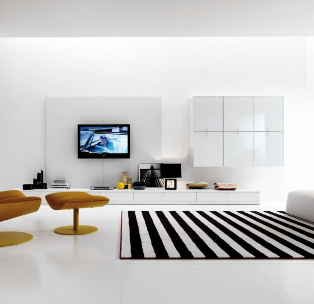 Modern minimalist living room with a white color palette, mustard-yellow lounge chair, black-and-white striped rug, and a built-in bookshelf filled with colorful books.