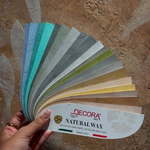 A hand holding a color swatch palette featuring 20 shades of Decora A&C Natural Wax, showcasing Italian-inspired luxury paints in soothing colors.