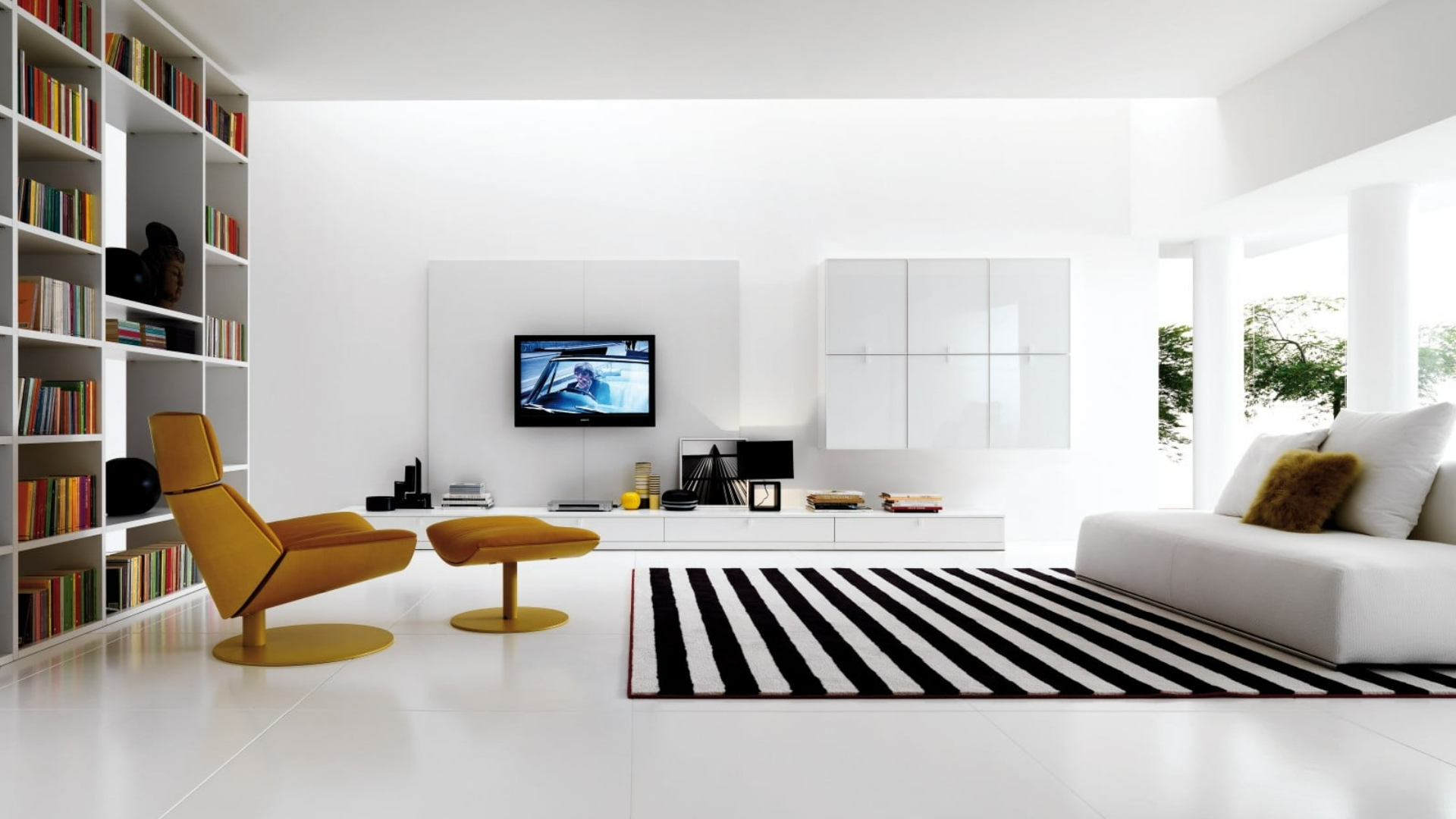 Modern minimalist living room with a white color palette, mustard-yellow lounge chair, black-and-white striped rug, and a built-in bookshelf filled with colorful books.